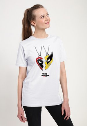 Junge Frau trägt ein weißes T-Shirt mit roten und gelben herzförmigen Superheldenmasken-Anhängern und dem Text „Come Together Marvel Studios.“
