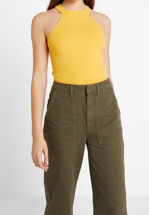 Vrouw draagt een mouwloze geel geribde haltertop en hooggetailleerde olijfgroene broek met voorzakken tegen een witte achtergrond.