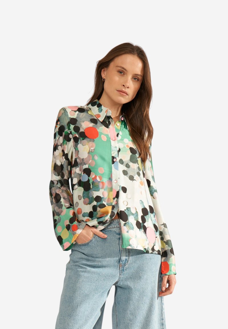 Femme portant un chemisier multicolore à pois et un jean bleu clair, debout avec une main dans la poche devant un fond uni.