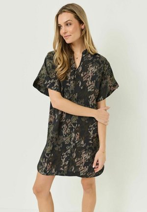 Vrouw met lang blond haar, gekleed in een loszittende korte jurk met een donker bloemenpatroon, staand tegen een lichte achtergrond, haar armen zachtjes gekruist.