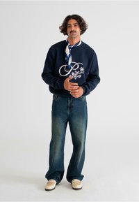 Marineblauer Pullover mit floraler Stickerei, kombiniert mit weit geschnittenen, blassen blauen Jeans und beigen Schuhen. Akzentuiert mit einem blau-weißen gemusterten Schal.