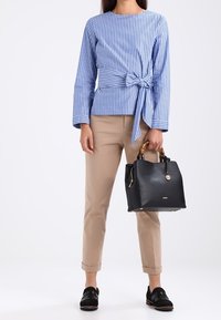 Žena oblečená v modro-bílém pruhovaném dlouhém rukávu s mašlí, béžových kalhotách, černých mokasínech, držící černou kabelku s bambusovými rukojetěmi.