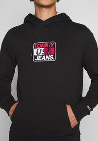 Zwarte hoodie van katoen met een voorvak. Bevat een rechthoekig logo in rood, wit en blauw met "TOMMY USA JEANS." op de borst.