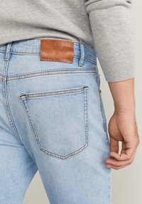 Jeans de mezclilla azul claro que presentan un parche de cuero en la parte posterior etiquetado "MNG MAN PREMIUM DENIM." Diseño de pierna recta con detalles de costura sutiles.