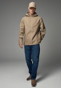 Beige wasserdichte Jacke mit Kapuze, Frontreißverschluss und Seitentaschen, kombiniert mit blauen Jeans und braunen Sneakern. Eine Schirmmütze rundet den Look ab.