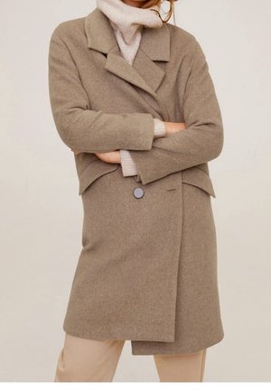 Femme portant un manteau en laine beige par-dessus un pull à col roulé crème, bras croisés, debout devant un fond clair uni.