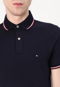 Marinblå polo med kontrasterande röd och vit randig krage. Har en tvåknappsknäppning och ett litet logopatch på bröstet.