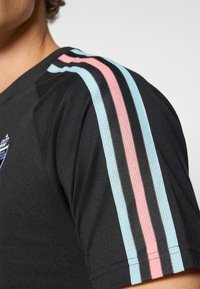 T-shirt de sport noir avec des rayures verticales roses et bleu clair sur les manches, fabriqué en tissu texturé et arborant un petit logo sur la poitrine.