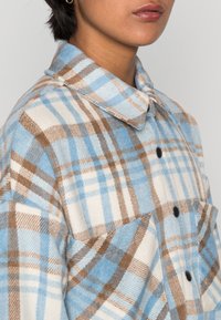 Camicia in flanella a quadri marrone e blu con colletto, caratterizzata da grandi bottoni, una tasca sul petto e una texture morbida.