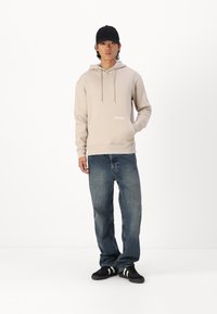 Felpa beige con cappuccio regolabile e tasca frontale, abbinata a jeans blu larghi e scarpe nere con strisce bianche.