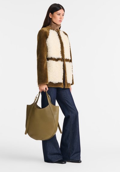 Woman wearing brown shearling jacket, brown turtleneck, dark flared jeans, black shoes, holding large olive green leather handbag.