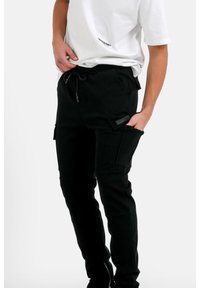 Zwarte cargo broeken met een taillekoord, zijzakken en een zachte, gestructureerde stof. G gedragen met een wit T-shirt, weergegeven tegen een effen achtergrond.