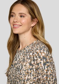 Bluse mit Rundhalsausschnitt, aus einem hellbeigen Stoff mit einem braunen und teal-farbenen abstrakten Muster. Strukturierte Falten an der Schulter.