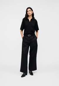 Μαύρο πόλο μπλούζα με φερμουάρ, συνδυασμένη με μαύρο φαρδύ παντελόνι, με ζώνη με ασημένια οβάλ αγκράφα και μαύρες μπότες με τακούνι.