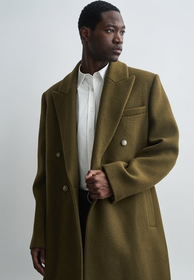 Vīrietis ģērbjas pārāk lielā olīvu zaļā dubultajā jaciņā virs baltās pogājām krekla, skatoties uz sāniem pret vienkāršu fona.