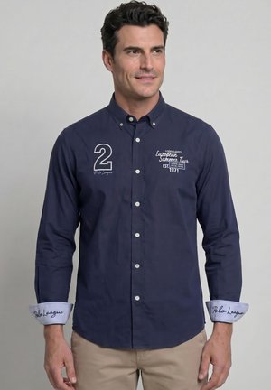 Hombre con camisa azul marino abotonada con texto blanco y número 2, pantalones beige y puños remangados con la inscripción "Polo League."