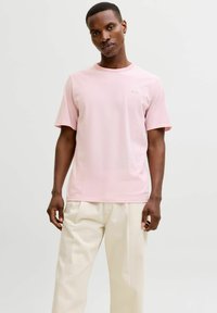 Hellrosa T-Shirt mit kurzen Ärmeln und rundem Ausschnitt, versehen mit einem dezenten Logo-Detail. Der Stoff wirkt weich und hat eine glatte Textur.