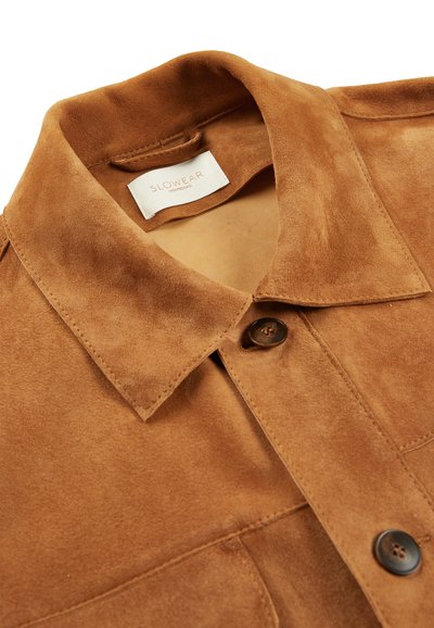 Veste en suede marron avec un col classique, fermeture à boutons sur le devant et détails de couture. Étiquette "SLOWEAR" sous le col.