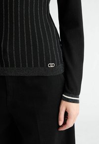 Zwart aansluitend top met verticale zilveren strepen, geribde manchetten met witte accenten en een logo-detail nabij de zoom. Textiel met textuur.