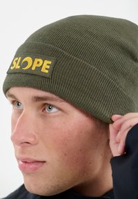 Dunkelgrüner Strickmütze mit strukturierter Oberfläche und einem gelben "SLOPE"-Logopatch an der Vorderseite. Nahaufnahme einer Person, die die Mütze anpasst.