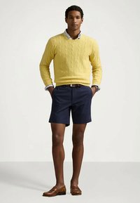 Camisola amarela de malha com padrão de tranças, calções azuis escuros e mocassins castanhos; modelo está com as mãos nos bolsos, vestindo uma camisa com colarinho por baixo da camisola.