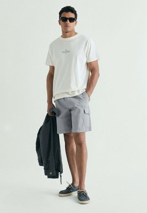 Hombre con gafas de sol, camiseta blanca, pantalones cortos cargo grises y zapatos náuticos azul marino, sosteniendo una chaqueta oscura con una mano en el bolsillo.