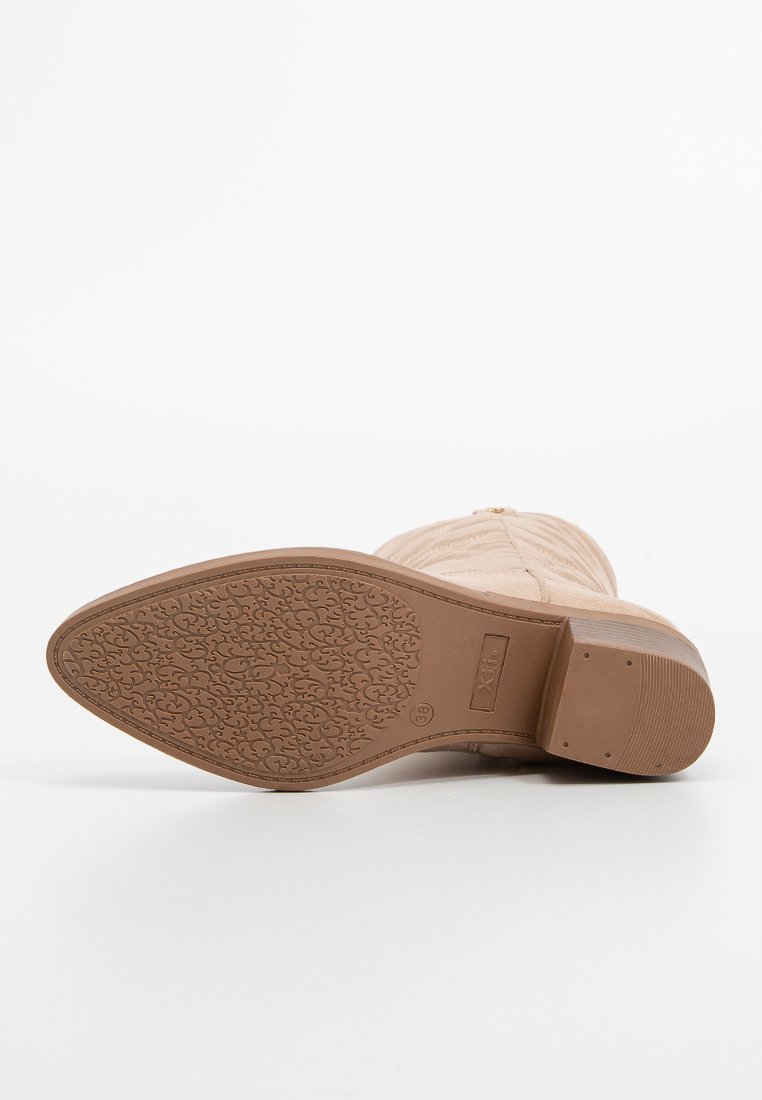 Bota de tobillo color beige claro con parte superior de sintético texturizado, tacón bajo en bloque, suela de goma con patrón y sutil costura decorativa.