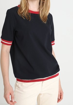 Kobieta ubrana w czarną koszulkę z krótkim rękawem z czerwono-białym wykończeniem na rękawach, kołnierzu i dolnej krawędzi, w połączeniu z jasnymi spodniami.