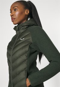 Dark green jacket with a quilted, padded front and textured knit sleeves. Features a high collar and a small logo on the chest.