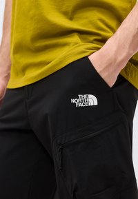 Άτομο με μαύρο παντελόνι The North Face με τσέπη με φερμουάρ, χέρι στην τσέπη και πράσινο πουκάμισο.