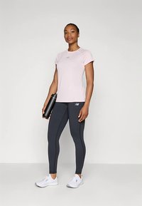 Žena stoji odjevena u svijetlo ružičastu majicu s kratkim rukavima, crne tajice, bijele tenisice, drži crnu bocu za vodu naslonjenu na jednostavnu pozadinu.