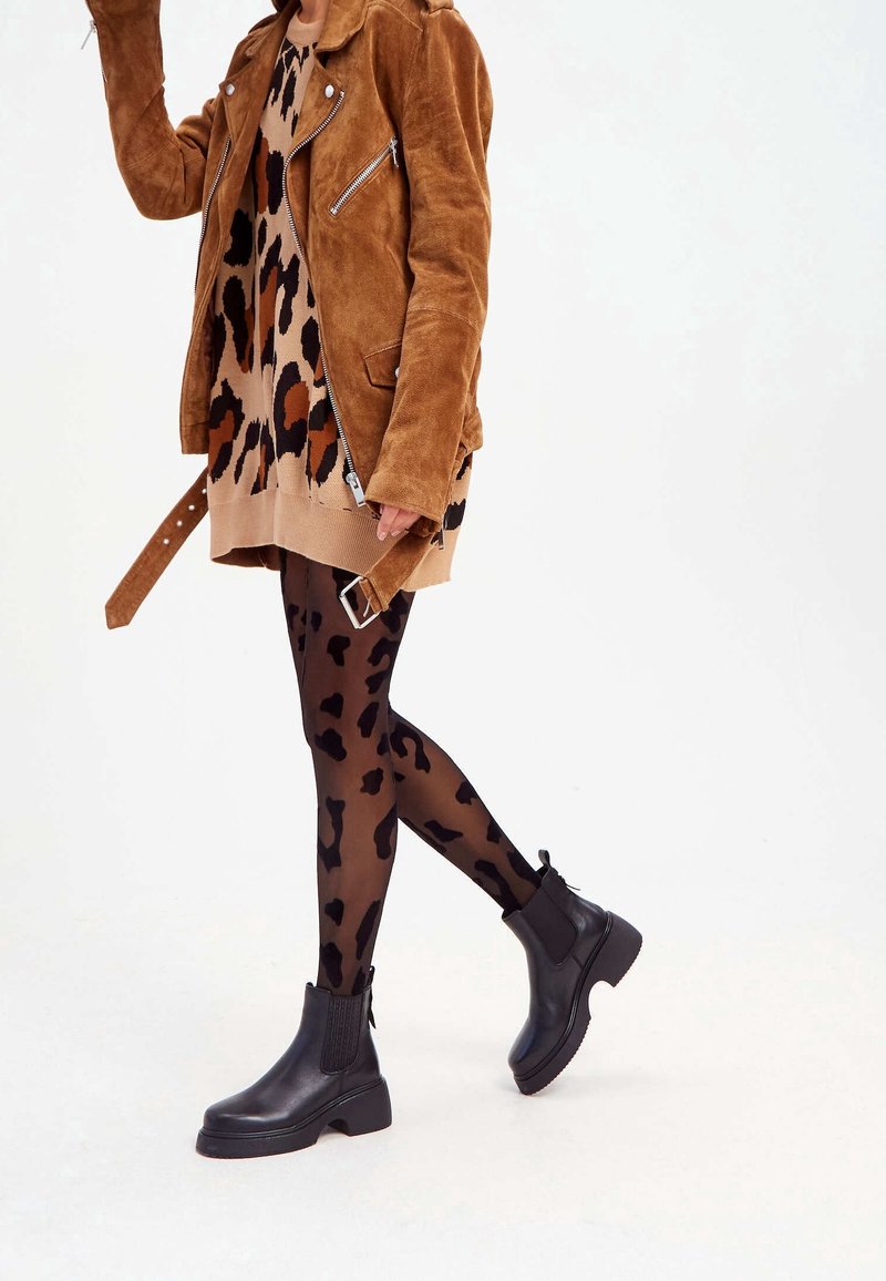 Braune Wildlederjacke über einem sandfarbenen Kleid mit Leopardenmuster, kombiniert mit schwarzen, leopardmusternden Strumpfhosen und klobigen schwarzen Stiefeletten.