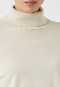 Zbliżenie osoby w kremowym swetrze z golfem o prążkowanej fakturze, ukazujące szyję i górną część klatki piersiowej.