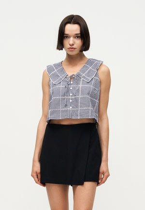 Νέα γυναίκα με ίσια σκούρα καρέ μαλλιά, που φοράει ένα μπλε καρό αμάνικο κοντό τοπ και μια μαύρη μίνι φούστα ψηλής μέσης.