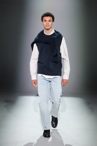 Pulover bleumarin aruncat peste o cămașă albă cu guler, blugi de culoare albastru deschis și pantofi negri purtați de un model care merge pe un podium gri.