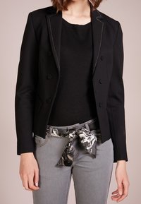 Černý dvouřadový blazer s kontrastním bílým prošitím, nošený přes černý top, v kombinaci se šedými džíny a šátkovým opaskem se vzorem.