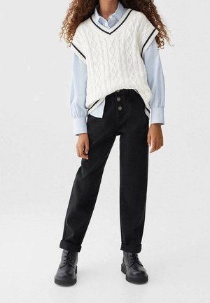 Žena ve světle modré košili, bílém pleteném vestě, černých džínách s vysokým pasem a černých šněrovacích botách stojící před jednobarevným pozadím.