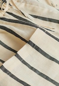 Gros plan sur un tissu beige avec des rayures horizontales noires et des coutures visibles, montrant une poche et un cordon de serrage noué.