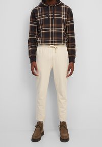Braun kariertes Hoodie mit Kordelzug, cremefarbene taillierte Hose und braune Wildlederstiefel. Das Design zeichnet sich durch einen strukturierten Stoff und kontrastierende Farben aus.