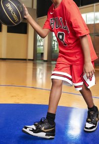Rode basketbalshirt en -shorts met zwarte accenten. Zwart-witte sneakers met gouden details. Houdt een basketbal vast, op een veld.