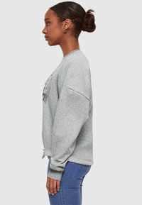 Grå sweatshirt med avslappnad passform, ribbade muddar och dragsko i nederkant. Har ett grafiskt tryck på framsidan och en mjuk textur. Matchas med blå jeans.