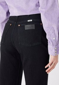 Czarne dżinsy wykonane z denimu mają wysoki stan, dwie tylne kieszenie oraz teksturowaną skórzaną naszywkę z logo "Wrangler" na pasku.