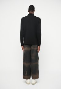 Pessoa de pé, voltada de costas, vestindo uma camisa preta de manga longa, calças largas preta e castanho com efeito tie-dye e bolsos, e ténis brancos.