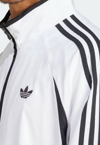 Chaqueta blanca de Adidas con rayas negras en las mangas, cremallera negra y logo trefoil de Adidas negro en el pecho, usada por una persona.