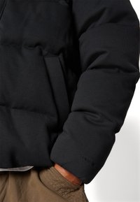 Jachetă pufă neagră cu textură matlasată, siluetă supradimensionată și broderie "THERM." Mână vizibilă în buzunarul pantalonilor de culoare maro deschis.