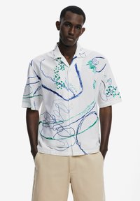 Camicia bianca a maniche corte con motivi astratti blu e verdi, chiusura con bottoni sul davanti e colletto da campeggio. Tessuto testurizzato con una vestibilità rilassata.