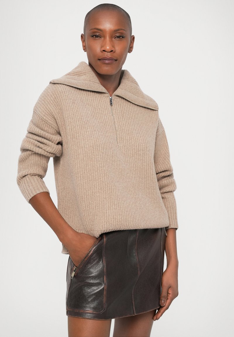 Kvinna med kort hår som bär en beige ribbad tröja med dragkedjekrage och en mörkbrun läderminikjol med dragkedjefickor, stående med en hand i fickan.