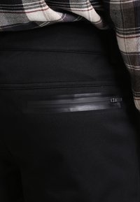 Czarne spodnie o gładkiej fakturze, z poziomą kieszenią na suwak z czarnymi elementami. Częściowo widoczna koszula w kratę powyżej.