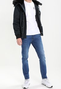 Svart pufferjacka med fleecefodrad huva, vit t-shirt, blå jeans och vita sneakers. Jackan har dragkedja och tryckknappslås.