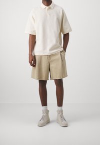 Světle béžové polo tričko s texturovaným vzorem, kombinované s khaki šortkami. Model má na sobě béžové vysoké tenisky.
